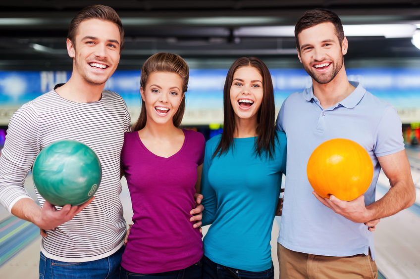 We love this game. Cheerful friends looking at camera and smiling while standing against bowling alleys